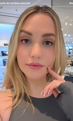 Blonde woman with shoulder-length hair, wearing a dark top, touching her face in a well-lit indoor setting.