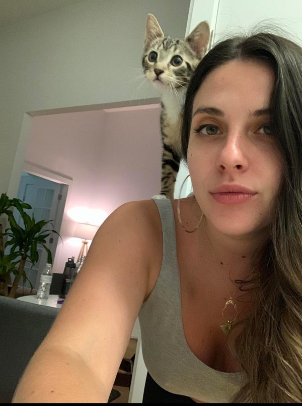 A woman with long dark hair takes a selfie in a gray tank top, with a tabby cat perched behind her shoulder in a cozy