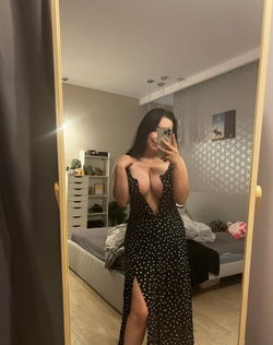 Brunette woman in a black polka-dot dress with a deep neckline takes a mirror selfie in a bedroom, partially revealing