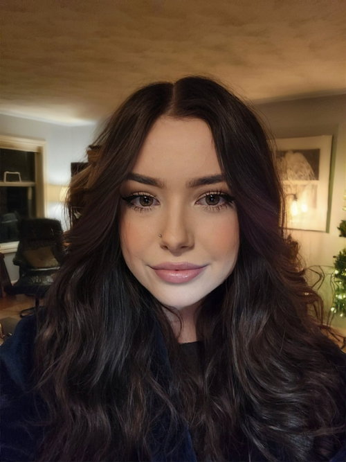 Brunette woman with long wavy hair and makeup in dark top in cozy indoor room.