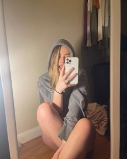 Blonde woman in a gray hoodie takes a mirror selfie, sitting bare-legged on the floor with her phone covering part of