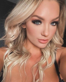 Blonde woman with wavy hair and makeup, wearing a black top, taking a close-up selfie indoors.