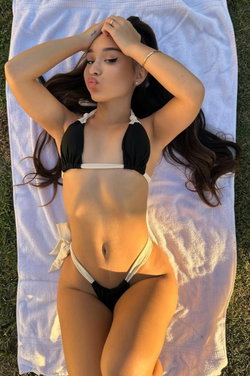 Brunette woman in black and white bikini lying on a towel outdoors, hands behind head, making a playful face.