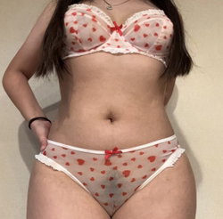 Woman with dark hair wearing sheer red heart-patterned lingerie with lace trim, standing against a plain wall.