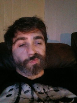 A man with dark hair and a beard, wearing a black graphic t-shirt, reclines on a dark couch indoors.