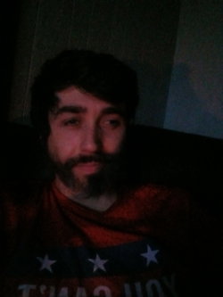 Man with dark hair and beard wearing a red graphic t-shirt, sitting in a dimly lit room.