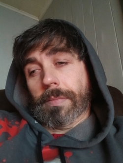 Man with dark hair and beard wearing a gray hoodie, looking directly at the camera in an indoor setting.