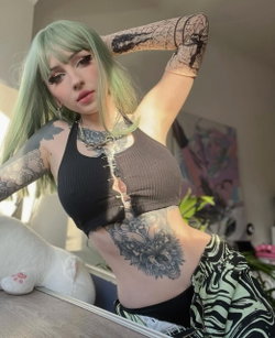 A person with green hair and tattoos poses in a black top and patterned pants.