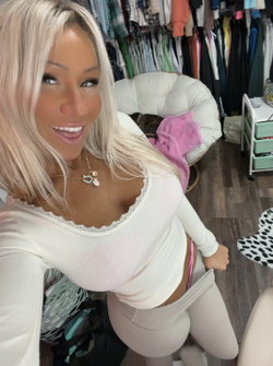Blonde woman in a white lace-trim top and beige leggings poses in a closet, pulling down her pants slightly.