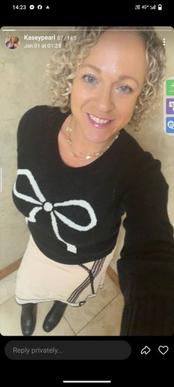 A woman with curly blonde hair smiles in a black sweater with a white bow design, paired with a light skirt and black
