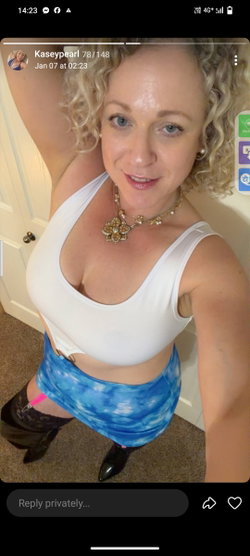 Blonde woman with curly hair in a white crop top and blue tie-dye skirt, posing indoors in a selfie-style shot.