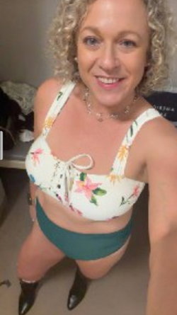 Curvy woman with curly blonde hair wearing a floral top and green bottoms, smiling in a selfie pose indoors.