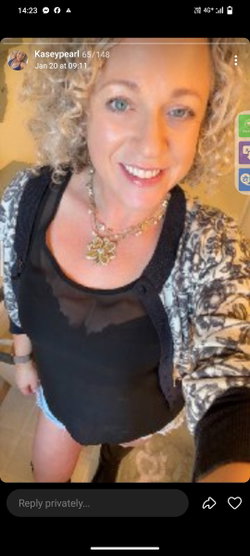 A smiling woman with curly blonde hair wearing a black top, patterned cardigan, and layered necklaces, posing indoors