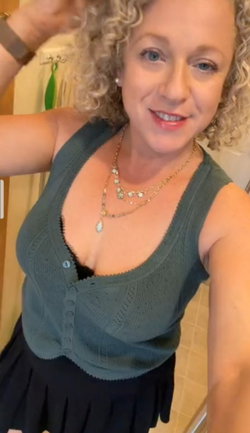 Curvy woman with curly blonde hair smiles in a selfie, wearing a green knit tank over a black top and layered necklaces