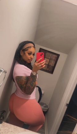 Curvy woman with tattooed arms takes mirror selfie in pink top and peach leggings, standing in bathroom.