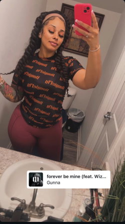 Curvy woman with long braids takes mirror selfie in bathroom, wearing a black patterned top and maroon leggings.