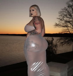 Curvy woman with blonde bob and tattoos wears sheer white dress, posing by a lake at sunset.