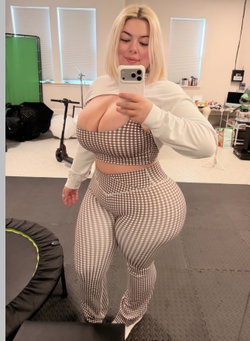 Blonde woman in a gingham two-piece set takes a mirror selfie in a home gym, wearing a white long-sleeve shirt pulled