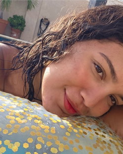 Close-up selfie of a woman with wet brown hair resting her head on a blue and gold patterned float in a pool, smiling