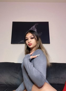 A woman with long ombre hair sits on a dark couch wearing a tight gray bodysuit, posing with one hand on her shoulder.