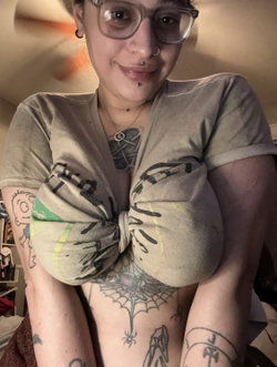 A woman with glasses and multiple tattoos wears a tied-up t-shirt, revealing chest and stomach tattoos in an indoor