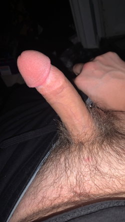 Close-up of a male genitalia with visible pubic hair, held by a hand, against a dark background with clothing.