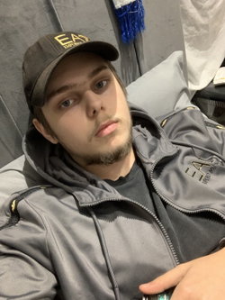 A young man with light eyes and a short beard wears a black cap and gray hoodie, lying on a couch in a casual indoor