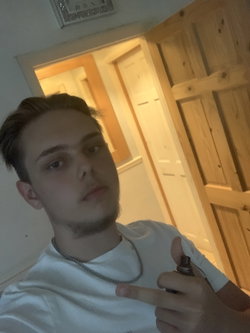 Young man with short dark hair and light stubble wearing a white t-shirt and chain necklace, holding a vape pen indoors
