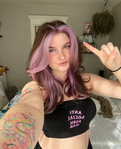 A woman with purple-tipped hair and colorful arm tattoos takes a selfie in a black crop top, posing in a bedroom with