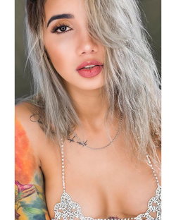 Woman with silver hair and colorful tattoo on shoulder, wearing a bejeweled top, posing closely for the camera.