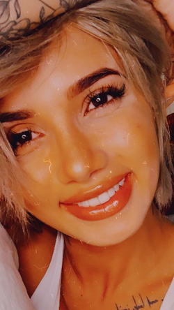 Close-up of a smiling woman with blonde hair, wearing a white top, with visible tattoos on her neck and hand.