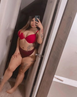 Curvy woman in red lingerie takes mirror selfie, showing tattoos on arms and legs, standing barefoot in a doorway.