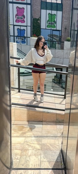 A woman with long dark hair takes a mirror selfie in a white crop top, maroon shorts, and a light jacket, standing on a
