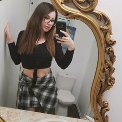 Woman with long brown hair and glasses in a black crop top and plaid skirt taking a selfie in front of a gold-framed
