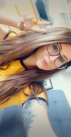 Woman with long brown hair, glasses, and a tattoo poses in a yellow top.