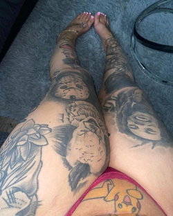Close-up of tattooed legs with intricate designs, wearing red lace underwear, resting on a plush gray surface.