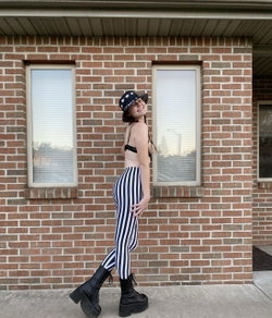 A woman with a curvy figure wears a black bra, striped leggings, and platform boots, posing outdoors against a brick