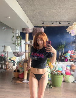 Redhead woman in black thong and t-shirt taking a selfie in a room with stuffed animals and plants.