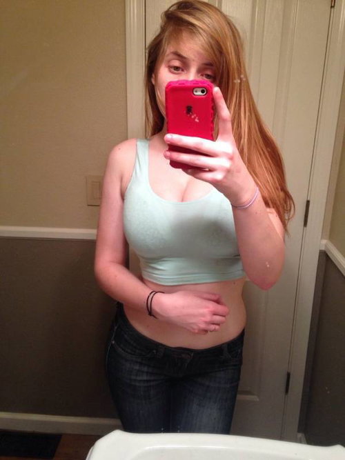A woman with long reddish hair takes a mirror selfie in a light blue crop top and dark jeans, holding a red phone.