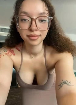 Curly-haired woman with glasses and visible tattoos on her arms, wearing a low-cut tank top in an indoor setting.