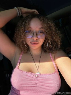 Curly-haired woman with glasses and hoop earrings wears a pink ruched top, posing indoors with one hand in her hair.