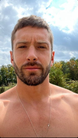 Shirtless man with short brown hair and beard, wearing a thin necklace, outdoors under a cloudy sky with trees in the