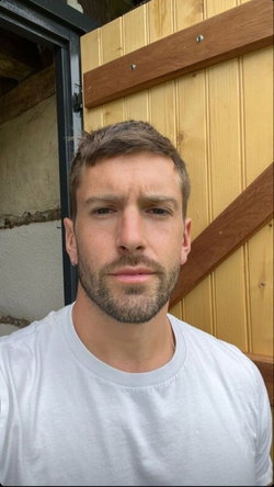 Man with short brown hair and beard wearing a white t-shirt, standing in front of a wooden door.