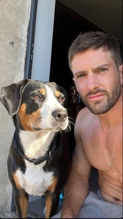 Shirtless man with short brown hair and beard poses with a black, white, and tan dog on a porch.