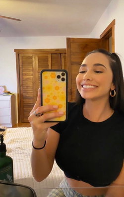 A smiling woman with dark hair takes a mirror selfie in a bedroom, wearing a black crop top and plaid bottoms.