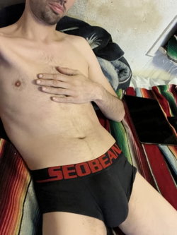 Shirtless man with hairy chest reclines on blankets wearing black boxer briefs.