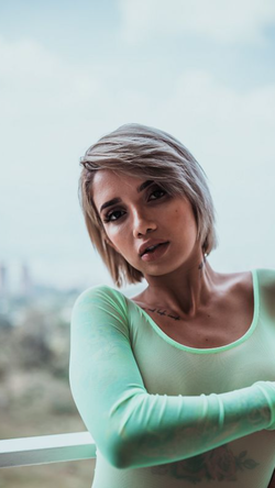 Woman with short blonde hair in a green top leaning on a railing with a cityscape in the background.