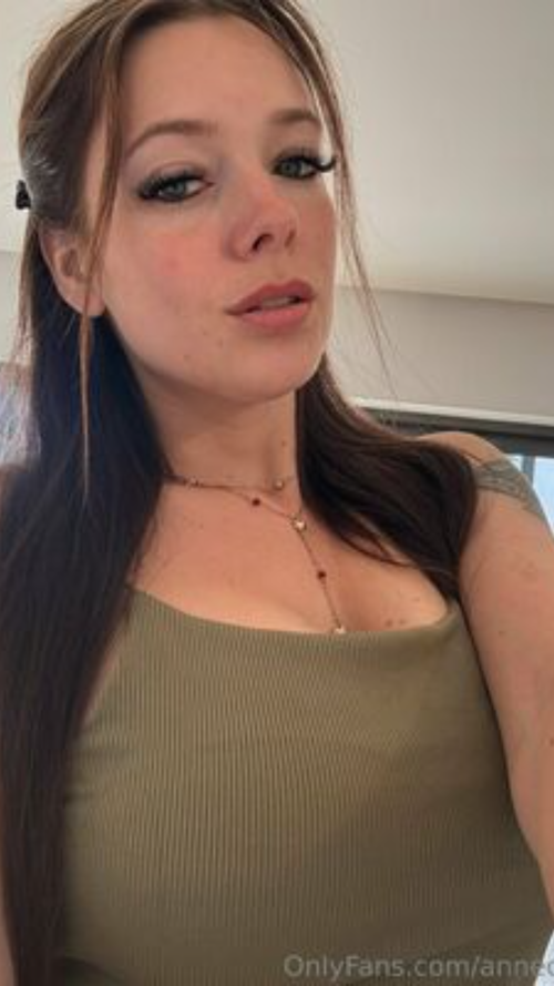 Brunette woman with long hair in a green ribbed tank top, wearing a delicate necklace, taking a close-up selfie indoors.