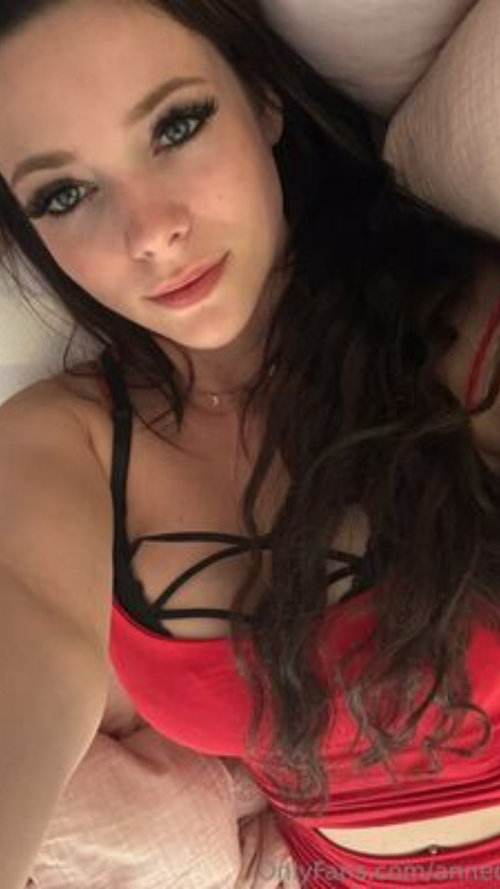 Brunette woman with long dark hair lying on a bed, wearing a red and black strappy top, looking directly at the camera.