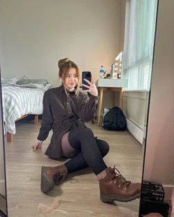 Blonde woman in a brown coat and black tights sits on the floor taking a selfie in a bedroom.